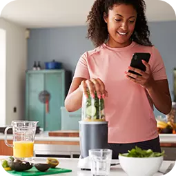woman making smoothie while smiling at phone